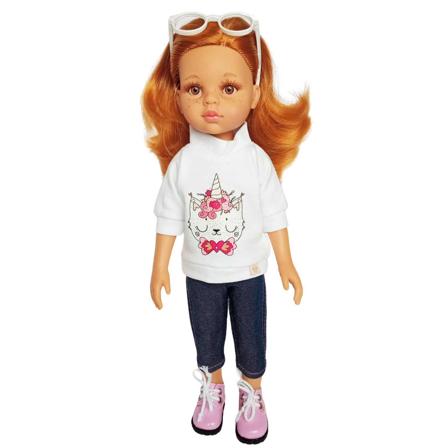 Paola Reina 32 cm doll wearing a white blouse with a unicorn kitty print, denim-style capris, pink lace-up shoes, and white sunglasses.
