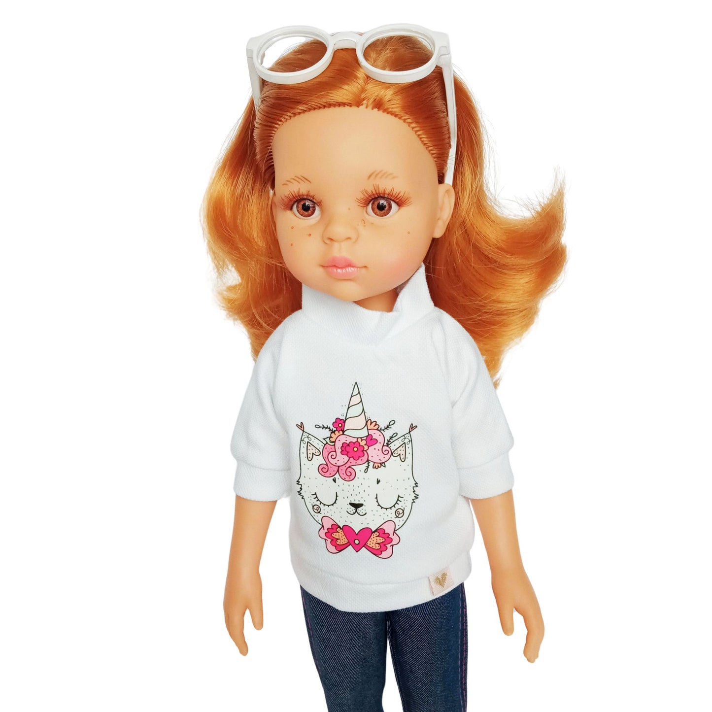 Paola Reina 32 cm doll wearing a white blouse with a unicorn kitty print, denim-style capris, pink lace-up shoes, and white sunglasses.